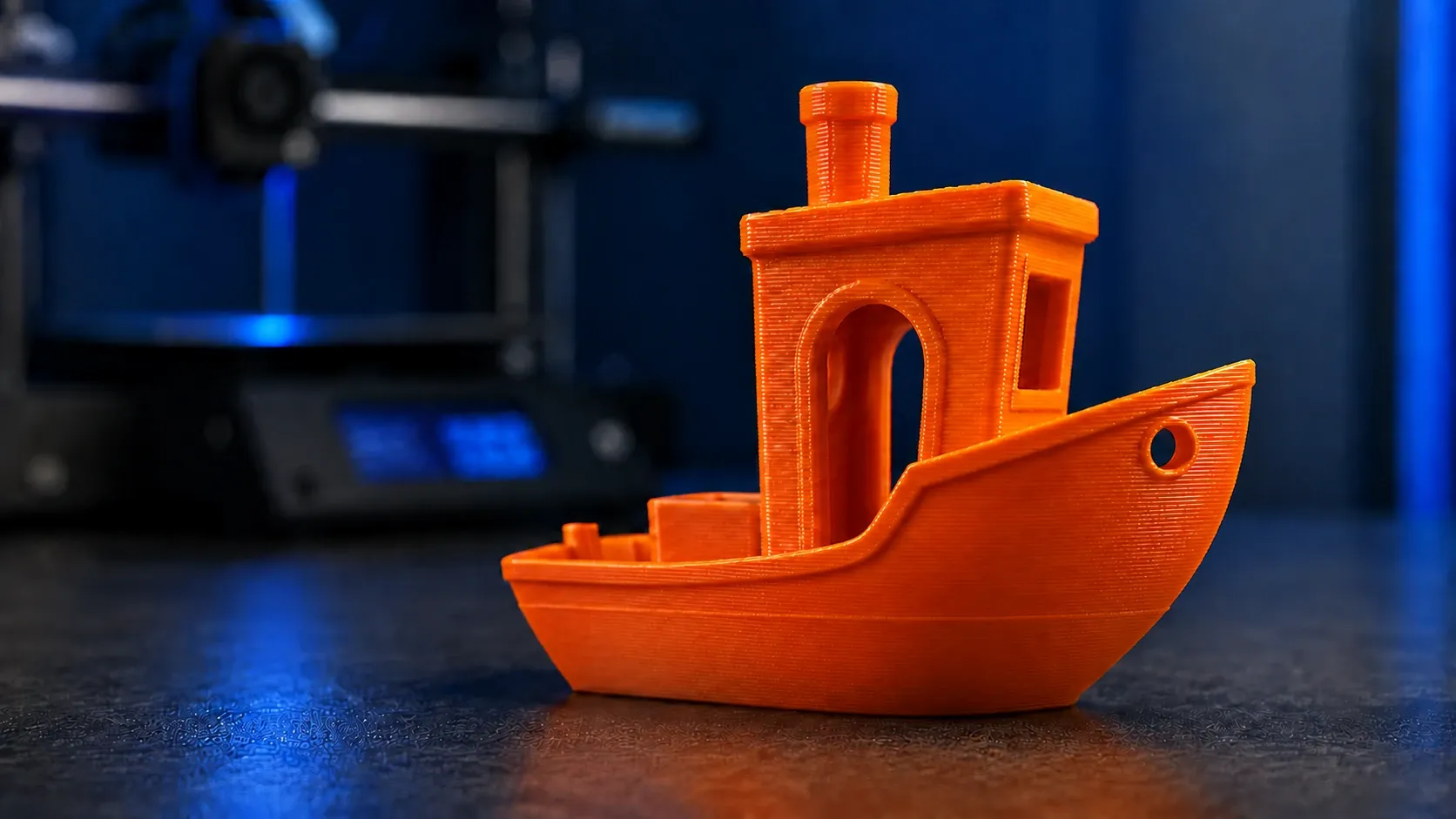 Ein orange gedrucktes Benchy-Schiffchen vor einem 3D-Drucker, die einzelnen Schichten sind im Streiflicht zu erkennen